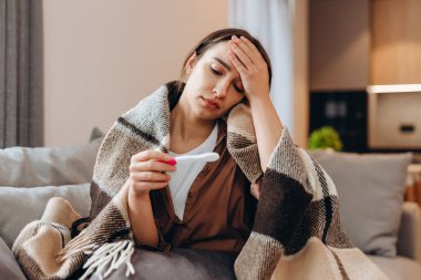 Sick sad girl sitting on the sofa covered with a blanket. He looks at her temperature.