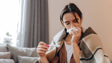 A sick woman measures her temperature while sitting on the couch and covering herself with a blanket.