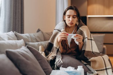 A sick woman measures her temperature while sitting on the couch and covering herself with a blanket