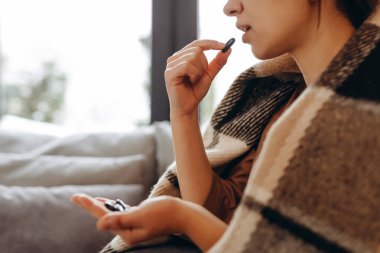 Young brunette woman in sweater, plaid, hold medicine, aspirin tablets, at home. Healthy lifestyle sick disease treatment cold season concept.