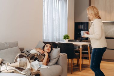A sick woman covered with a blanket, another woman brings a cup of tea.
