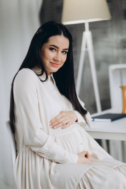 Hamile kadın gülümsüyor, kameraya bakıyor ve klinikte bir şey gösteriyor..