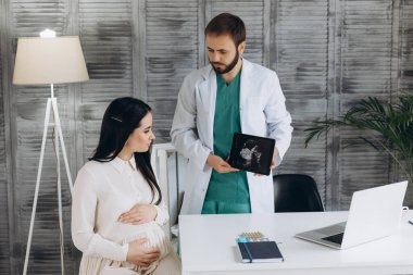 Doktor hamile bir kızın ultrasonunu gösteriyor. Klinikteki doktora geldi. Bir jinekolog. Beyaz bir bornoz giymiş..