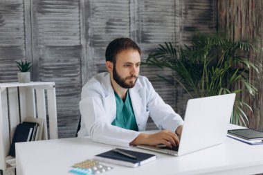 Çekici genç erkek doktor ışık dolabında dizüstü bilgisayarla çalışıyor. Gülümsüyor ve kameraya bakıyor..