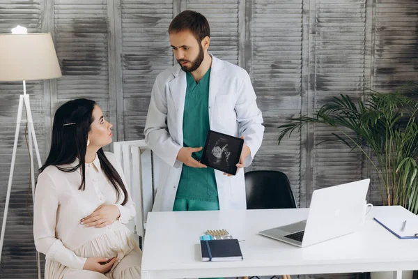 Stock image Doctor showing a pregnant girl picture ultrasound. She came to the doctor in the clinic. It's a gynecologist. He is dressed in a white robe.