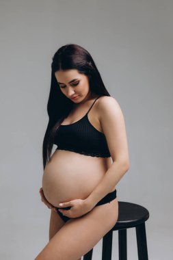 Hamile kadın karnına dokunuyor ve gri arka planda poz veriyor.