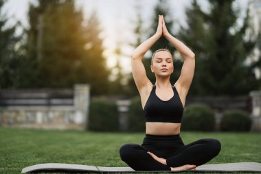 Spor kıyafetli genç bir kadın parkta yoga yapıyor. Sağlıklı bir kadın sabah lotus pozuyla çimlerde ve meditasyonda oturuyor. Sağlık, fitness, egzersiz ve iş hayatı dengesi kavramları.