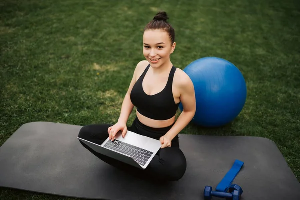 一个微笑的年轻健身女孩的肖像坐在户外垫子上使用膝上型电脑 — 图库照片
