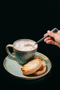 Kapuçino, kremalı çorba, kızarmış ekmek ve siyah arka planda kaşık.