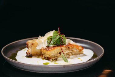 Krema soslu lazanya, yeşilliklerle süslenmiş ve siyah arka planda yeşil yağ var.