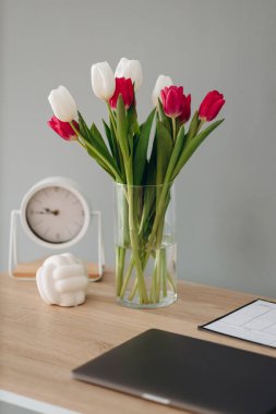 Masaüstünde cam vazodaki beyaz ve kırmızı laleler. Gri bir kara zeminde saat, dizüstü bilgisayar ve planör.
