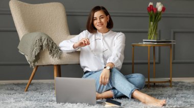 Ciddi bir iş kadını dizüstü bilgisayarın önünde yerde gri bir halıda oturuyor, beyaz gömlek ve açık kot pantolon giyiyor, elinde gözlük tutuyor, bej rengi bir koltuğa yaslanmış gri bir ekose ve içinde lale vazosu olan bir kahve masası olan bir vazo.