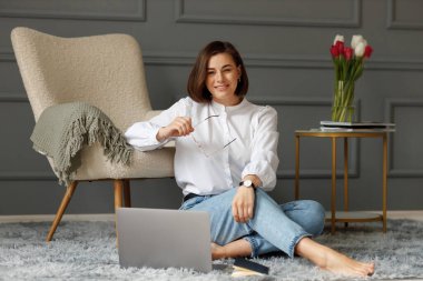 Ciddi bir iş kadını dizüstü bilgisayarın önünde yerde gri bir halıda oturuyor, beyaz gömlek ve açık kot pantolon giyiyor, elinde gözlük tutuyor, bej rengi bir koltuğa yaslanmış gri bir ekoseli ve üzerinde lale vazosu olan bir kahve masası ve bir vazo dolusu... 