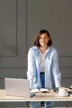 Önüne bakan, beyaz tişört, mavi gömlek ve açık renk kot giyen iş kadını masanın üzerine dizüstü bilgisayar ve ofis malzemeleriyle yaslanıyor.