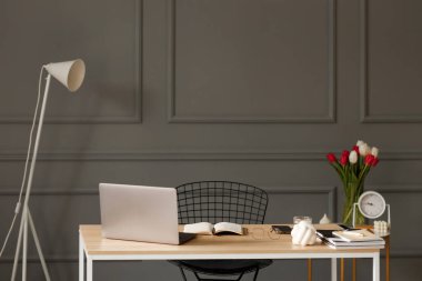 Bir defter, bir kalem, bir dizüstü bilgisayar, bir bardak su, yanında beyaz bir lamba, arkasında tablet, bir vazo, bir çiçek ve dekore edilmiş gri bir duvarda masa saati olan bir sandalye ve bir sehpa olan ahşap masa. 