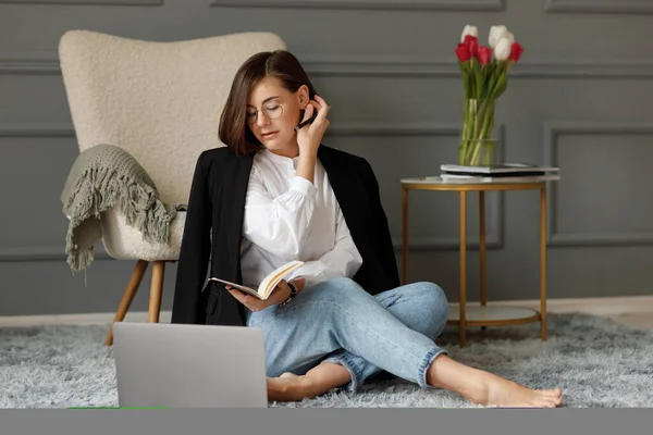 Businesswoman is sitting on a gray carpet on the floor in front of a laptop, is wearing a white shirt with a black jacket on her shoulders and light jeans, holds a pen in her hand and fix her hair behind her ear, is holding a notebook in the other ha