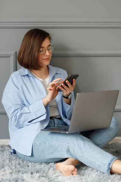 A business lady wearing a white t-shirt, blue shirt and light jeans is sitting on a gray carpet on the floor, leaning against a decorated gray wall, scrolling through her phone, holding a laptop on her legs