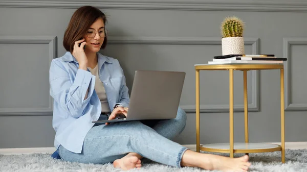 Business lady wearing a white t-shirt, blue shirt and light jeans sitting on a gray carpet on the floor near a table with a cactus vase and notebooks, leaning against a decorated gray wall, is talking on the phone, holding a laptop on her legs