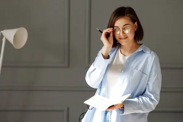 Business lady wearing a white t-shirt, blue shirt and light jeans, is standing on a gray background with a white lamp, holding a pen and glasses on her eyes in one hand and a notebook in the other hand in an office