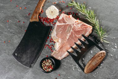 Siyah bir tabağa tuz, kırmızı ve siyah biber serpiştirilmiş kemikli biftek, biberiye ve yarım sarımsak serpiştirilmiş gri taştan kesme tahtasının yanında büyük mutfak bıçakları.