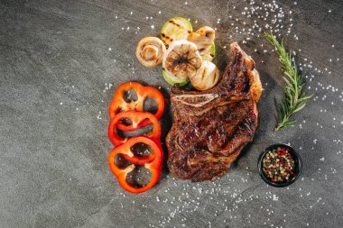 Koyu metal kesim tahtasının üzerinde sulu bir biftek, aromatik biberiye filizi, siyah bir biber karışımı, üç dilim taze kırmızı biber, ızgara mantar ve profesyonel bir mutfaktaki taş bir masanın üzerine tuz serpiştirilmiş kabak.