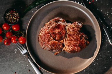 Büyük bir parça kurutulmuş biftek, yanında çatal ve bıçak, yeşil ve kırmızı biber, bir dal kiraz domatesleri, masaya tuz ve biber serpiştirilmiş siyah bir tencere karışık biber serpiştirilmiş, düz gri bir tabakta yatıyor.