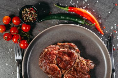 Düz gri bir tabakta duran ızgara bifteğin bir parçası, yanında çatal ve bıçak, yeşil ve kırmızı biberler, bir dal kiraz domatesleri, masada bir kavanoz karışık karabiber.