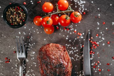 Masada çatal, bıçak, yeşil ve kırmızı biberli ızgara biftek, bir tutam vişneli domates, tuzlu ve biberli bir masada karabiber.