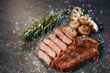 Pişmiş ve dilimlenmiş ızgara sulu et parçası tuz ve bezelye serpintisinin üzerinde, biberiye filizi yanında oturuyor ve profesyonel bir mutfakta taş bir masada mantar pişiriyor.