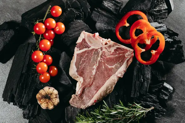 Juicy beef steak, with a sprig of rosemary, a sprig of cherry tomatoes, round slices of red sweet pepper and sliced roasted garlic on a charred log on a stone table