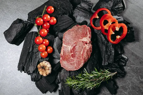 Juicy steak, with a sprig of rosemary, a sprig of cherry tomatoes, round slices of red sweet pepper and sliced roasted garlic on a charred log on a stone table on a kitchen