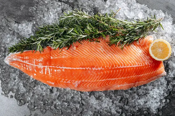 stock image Juicy raw red salmon steak, carefully cut lengthwise, on a bed of ice with sprigs of aromatic rosemary and a slice of lemon