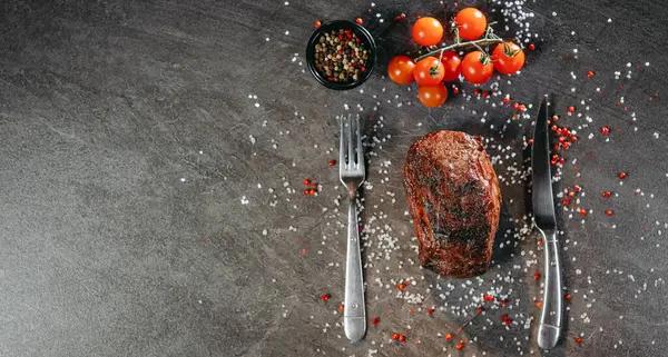 A generous piece of grilled steak on a table with fork and knife cutlery and green and red hot peppers, a sprig of cherry tomatoes, a black pot of mixed peppercorns on a table with salt and pepper