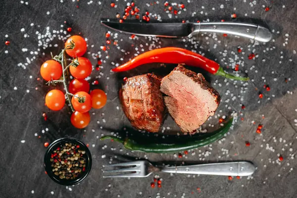 Sliced halved lean piece of seared steak on the table, next to which are cutlery fork and knife, as well as a sprig of cherry tomatoes, a black pot with a mix of peppercorns on the table, on which salt and pepper are scattered