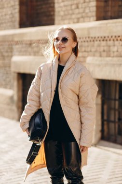 Gözlerinde güneş gözlüğü olan şık bir kız siyah bir süveter, siyah deri pantolon, yüksek siyah çizmeler ve bej bir yağmurluk giyiyor.