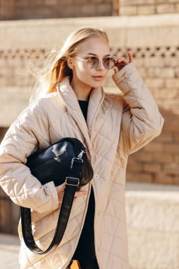 Gözlerinde güneş gözlüğü olan zarif bir bayan elinde siyah bir kazak, siyah deri pantolon, yüksek siyah çizmeler ve bej bir yağmurluk giyiyor. Eski bir binanın arka planına yakın bir çanta tutuyor.