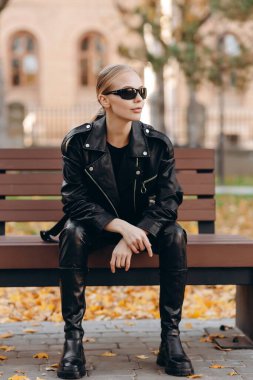 Gözlerinde siyah güneş gözlüğü olan zarif genç bir kadın, siyah bir tişört, siyah deri bir ceket, siyah deri pantolon ve uzun siyah botlar giyiyor, siyah deri çantasını yanında tutuyor ve kollarını bir bankın önünde çapraz oturarak oturuyor.
