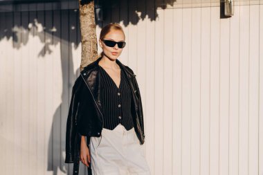 Saçları arkaya çekilmiş, siyah gözlüklü, siyah bir yelek giyen, omuzlarında siyah deri bir ceket ve beyaz geniş bir pantolon tutan ciddi bir kadın, ince bir ağaca yaslanmış, beyaz bir çitin arkasında duruyor.