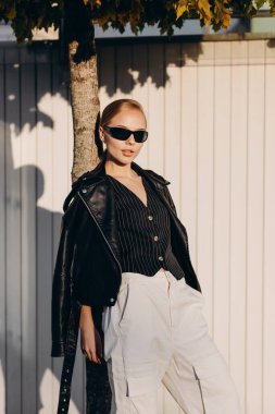 Saçı geriye çekilmiş, siyah gözlüklü, siyah bir yelek takmış, omuzlarında siyah deri bir ceket ve beyaz geniş bir pantolon tutan zarif bir kadın, ince bir ağaca yaslanmış, beyaz bir çitin arkasında duruyor.