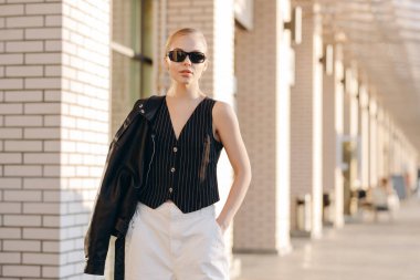 Siyah güneş gözlüklü, at kuyruklu güzel bir kadın, beyaz çizgili siyah bir yelek giyiyor, omuzunda deri bir ceket ve beyaz bir pantolon var, elini cebine sokmuş, arka planda bir B 'nin arka planında duruyor.