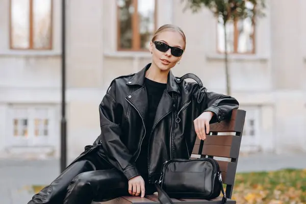 Stylish young woman with black sunglasses on her eyes, wearing a black t-shirt, a black leather jacket, black leather pants and tall black boots, holds her black leather bag near and sits on a park bench leaning on it and lookind at the camera agains