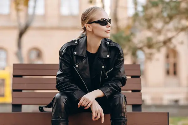 Serious fashionable woman with black sunglasses on her eyes looking away wearing a black t-shirt, black leather jacket and black leather pants sitting on a bench with her hands crossed on her knees against the background of a brick house