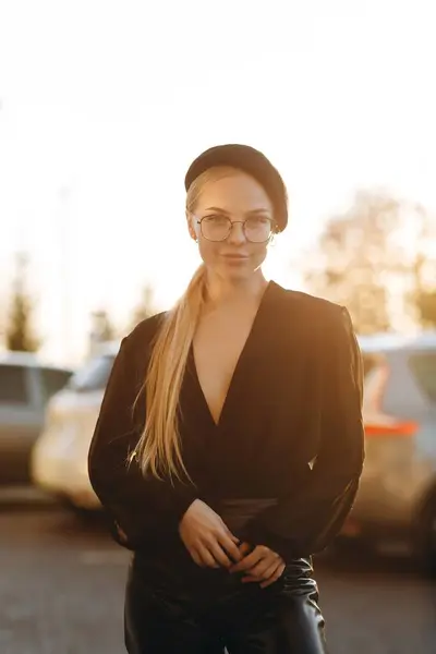 Elegant girl with light daytime make-up and pulled hair, with clear glasses on her eyes and a black beret on her head, looks right at the camera is holding her hands in front, wearing a black blouse, standing against the background of parked cars and