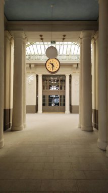 Sütunlarla dolu açık salonda saat tavandan sarkıyor