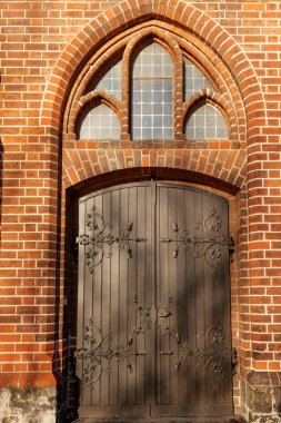 Berlin, Almanya - 15 Aralık 2019: Tarihi bir kilisenin gotik penceresinin altında işlenmiş demir menteşeli eski bir ahşap kapıyı aydınlatan güneş ışığı. Meryem Kilisesi