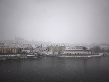 Prague, Czech republic - January 5, 2025: Snow gently falls over the historic riverside and castle, blanketing the charming architecture and creating a serene winter atmosphere clipart