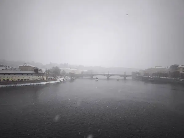 Prag, Çek Cumhuriyeti - 5 Ocak 2025: Kar, bulutlu bir gökyüzü altında sakin bir kış manzarası yaratan tarihi köprüler ve binalar ile huzurlu Vltava Nehri 'ni örtüyor