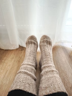 Pencere perdesinin yanında ayaklarını ısıtan yün çoraplar ve ahşap iç kısımları konfor ve sıcaklıkla dolduran ılık kış ışığı.