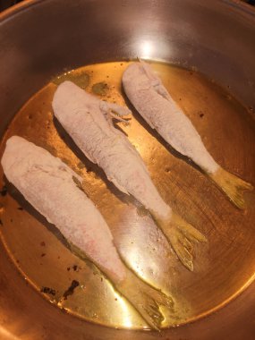 Üç küçük balık tavada zeytinyağında kızartılıyor, bir restoran mutfağında şef tarafından hazırlanıyor.