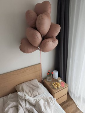 Kalp şeklinde moka köpüğü, gül rengi balonlar, taze kruvasanlarla kahvaltı seti, kahve, komodindeki kırmızı güller sakin bir yatak odasında romantik bir sevgililer günü sürprizi hazırlar.